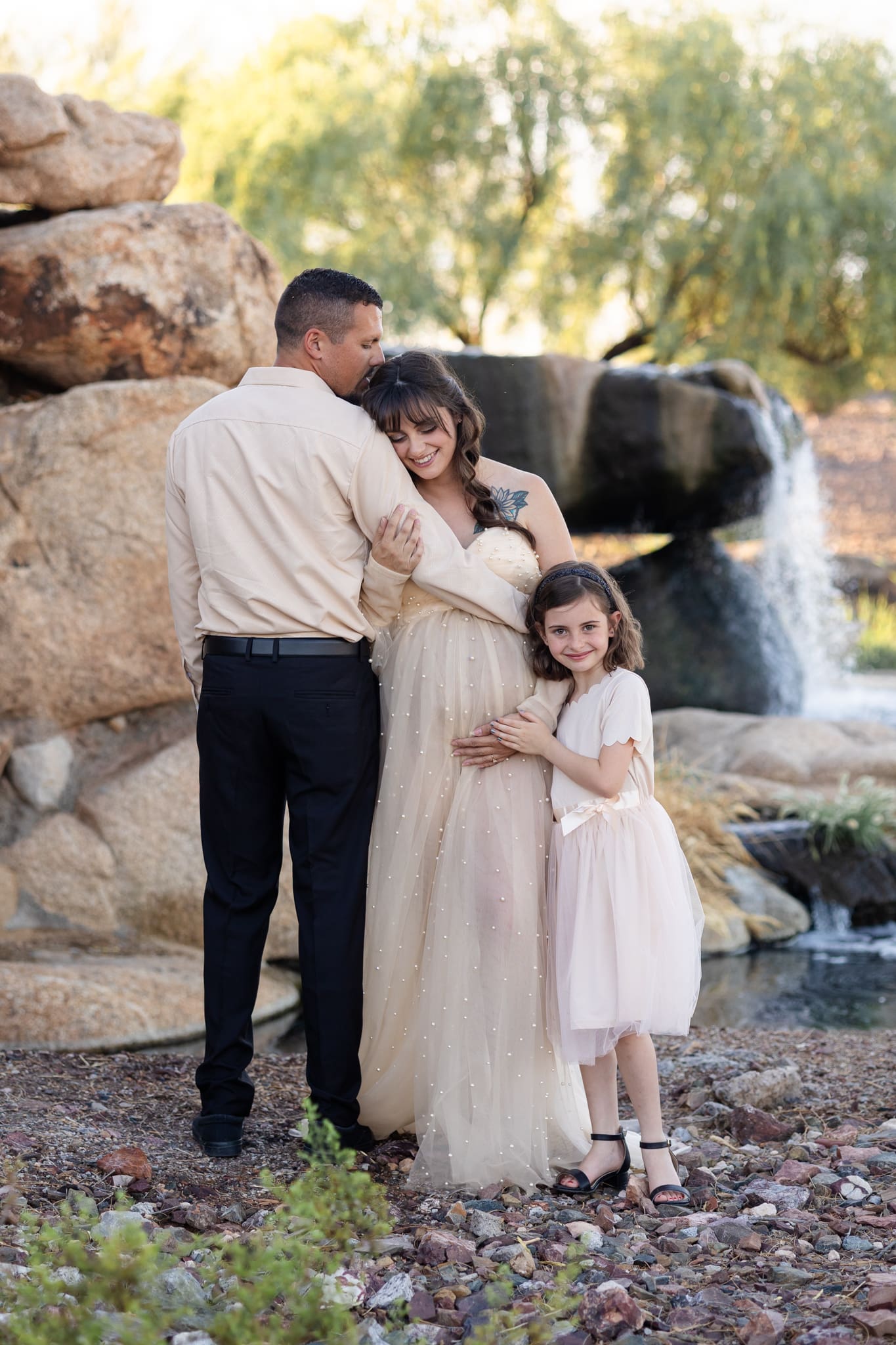 Pregnant woman in pearl-embellished gown embraces her partner while daughter hugs her baby bump by a waterfall.