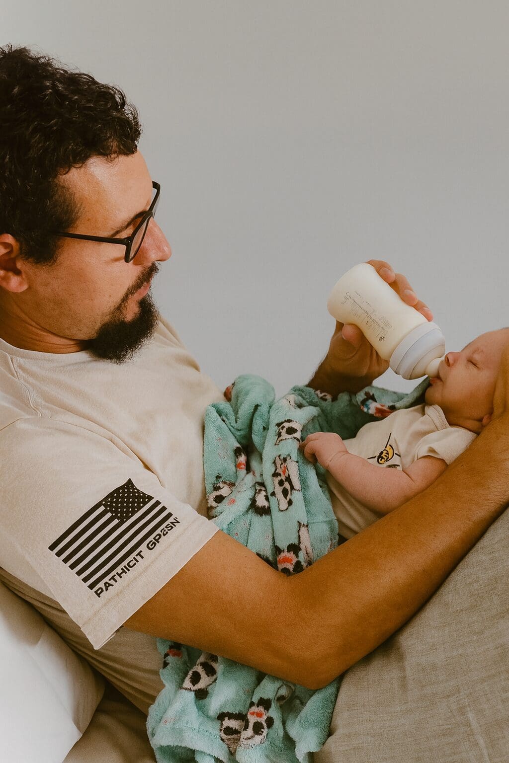 A father gently bottle-feeding his newborn baby while sitting on a bed, with a soft turquoise panda blanket wrapped around the infant.